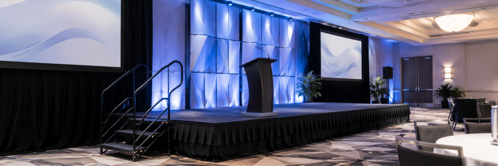 Indoor corporate stage rental in Orlando with black skirting, stairs, lectern, projection screens and blue event lighting.